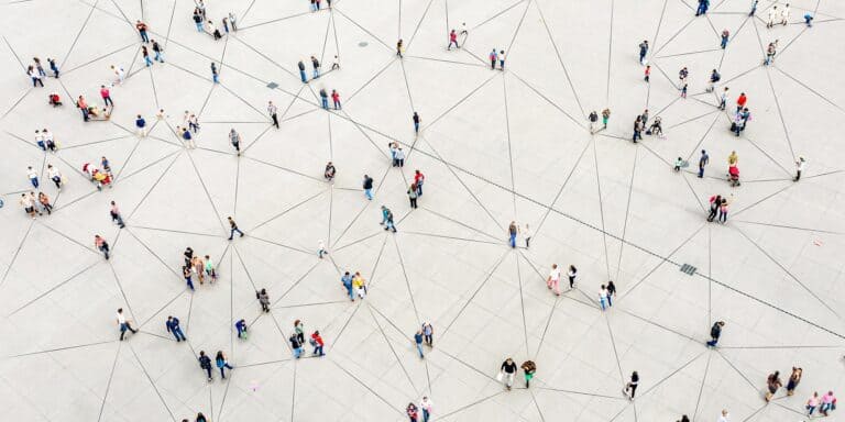 Aerial view of people walking on a large, light-colored open space with thin black lines connecting them in a web-like pattern. Individuals and small groups are scattered throughout, creating a visual network effect.