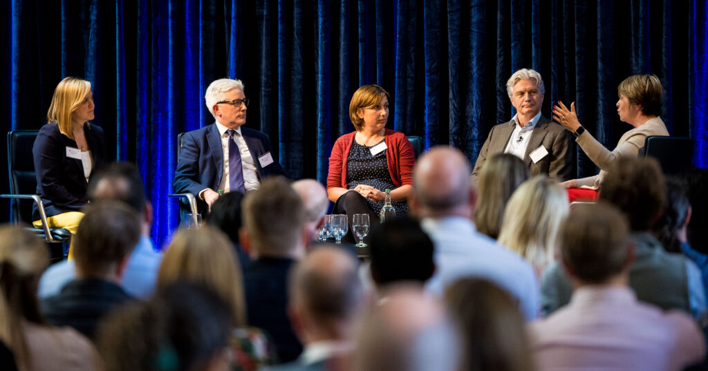 Five panelists sit on stage in front of a blue curtain, engaged in discussion, while an audience listens attentively. One panelist gestures while speaking.