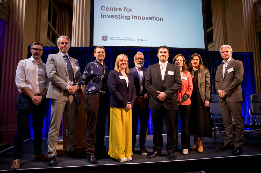Ten people in business attire stand on a stage under a screen that reads "Centre for Investing Innovation." They are posing for a group photo at a professional event in a large, well-lit hall.