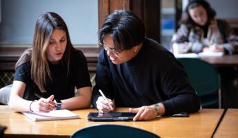 Two students sit at a table, collaborating and writing in notebooks and on a tablet, while another person studies in the background. They appear focused and engaged in their work.