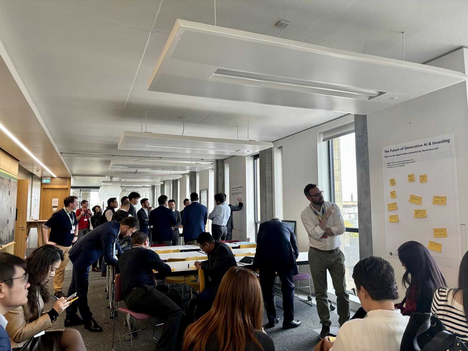 A group of people in business attire participate in a workshop, discussing and writing on large sheets of paper with sticky notes on the walls in a bright, modern conference room.
