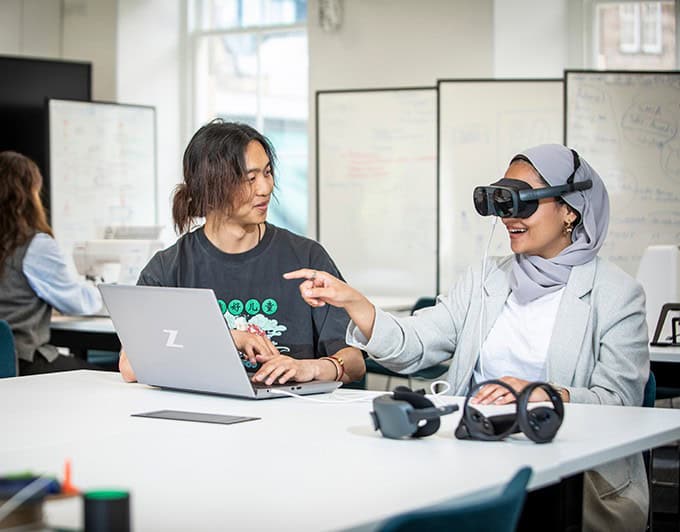 Two people sit at a desk; one types on a laptop while the other, wearing a VR headset, points at the screen. Whiteboards with diagrams and notes are visible in the background.