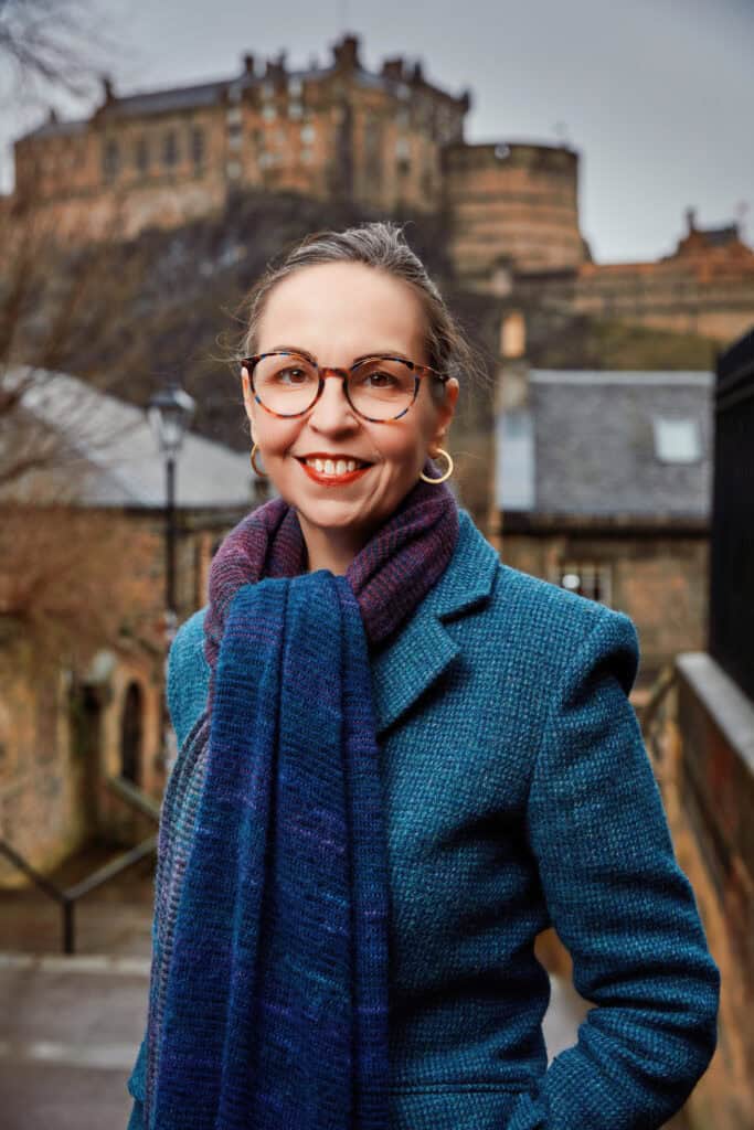 A smiling woman wearing glasses, a blue coat, and a scarf stands outdoors in front of a historic stone castle on a hill, with overcast skies and old stone buildings in the background.
