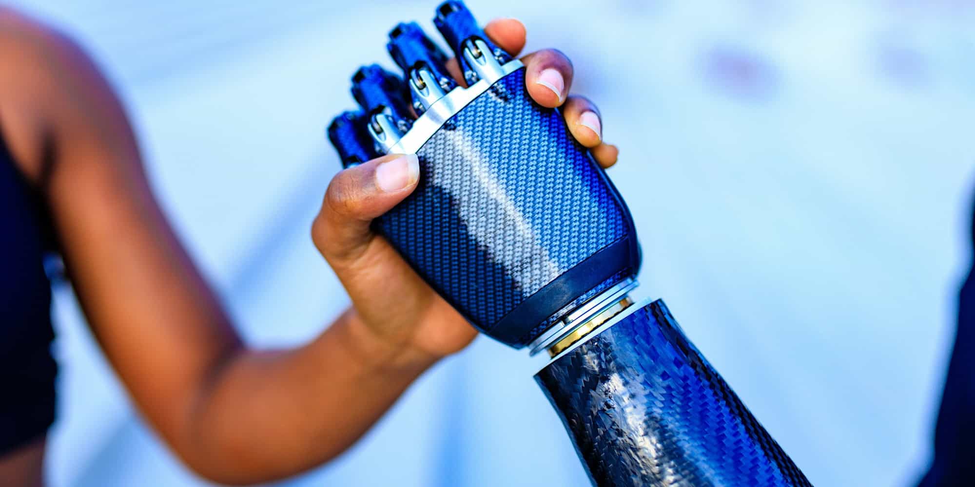 A close-up image of a person with dark skin shaking hands with a robotic arm. The person's hand is gripping the metallic blue and black robotic hand, capturing the connection between human and robotic technology. The background is blurred.