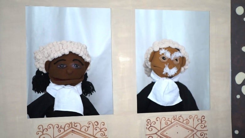 Two puppets wearing judge's wigs and black robes are displayed against a beige background. Each puppet is set in a separate window; the puppet on the left has dark skin and black hair, and the puppet on the right has lighter skin and white hair. Decorative designs are below them.