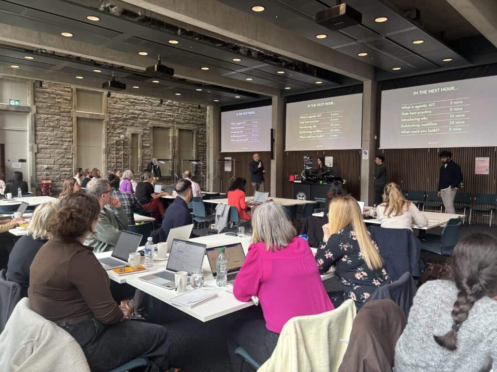 A group of people in a modern conference room listen to presenters at the front, where two large screens display an agenda. Attendees sit at tables with laptops, notebooks, and drinks.