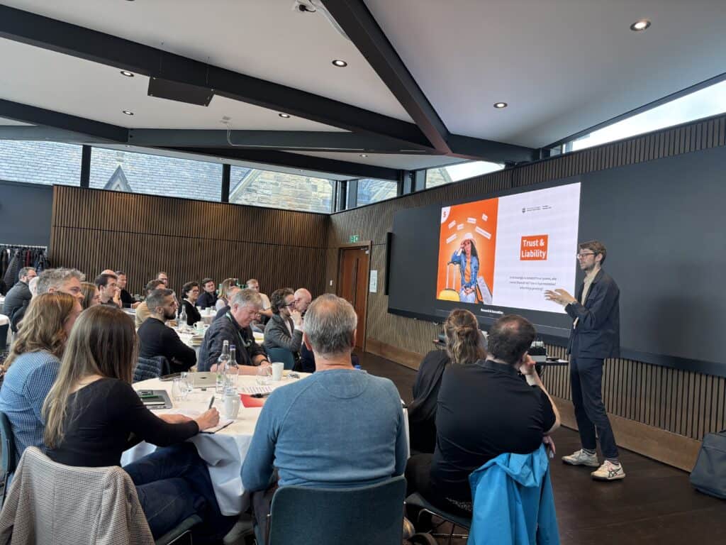 A person stands and presents to a group of people seated at tables in a modern conference room. A large screen displays a slide titled “Trust & Liability” with graphics and text.