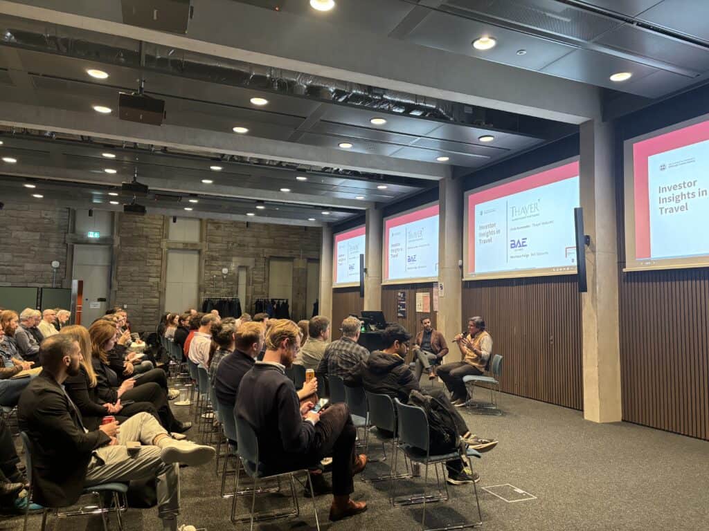A group of people sit in rows facing a panel discussion at a conference. Large screens display "Investor Insights in Travel" and logos. The room has modern decor with wood panelling and stone walls.