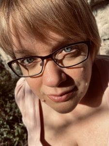 A person with short blond hair and glasses looks up at the camera, standing outdoors in sunlight with some shadows on their face.