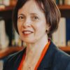 katharine-aulton A woman with short brown hair wearing a dark blazer and an orange top stands in front of a bookshelf, smiling slightly at the camera.