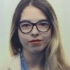 A young woman with long, light brown hair, wearing black-rimmed glasses, light red lipstick, a blue shirt, and a light-colored jacket, looks directly at the camera against a plain background.