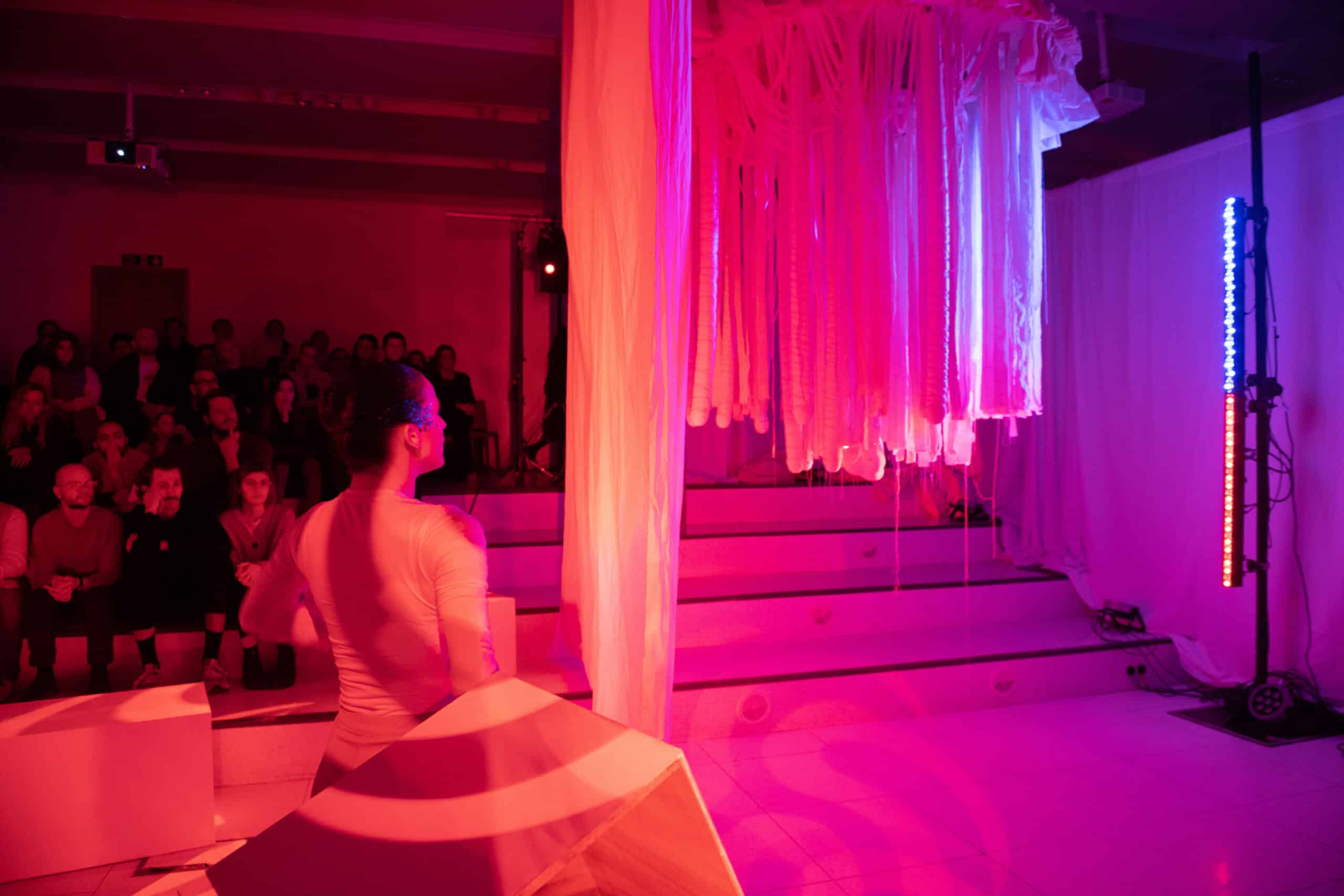 Dancer facing the hanging white tentacles, mood lighting orange, with an audience in the background