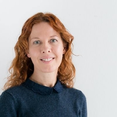 A woman with wavy, red hair is smiling at the camera. She is wearing a dark blue sweater with a delicate collar. The background is plain white.