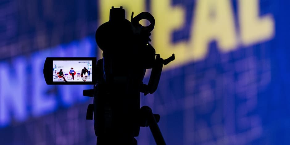 A video camera with a panel discussion visible in the viewfinder screen is silhouetted against the New Real logo.
