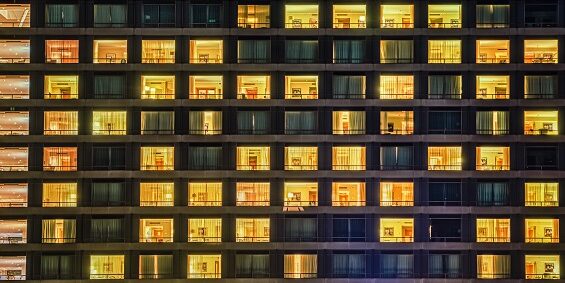 Building facade shows a windows and rooms pattern at night. The exterior features illuminated and dark living spaces with people living like neighbours in a shared communal space.