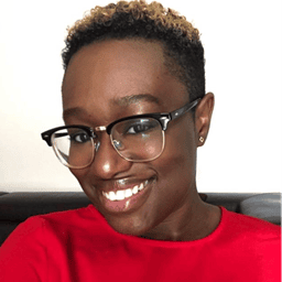 A person with short, curly hair and glasses is smiling at the camera. They are wearing a red top and are positioned against a plain background.