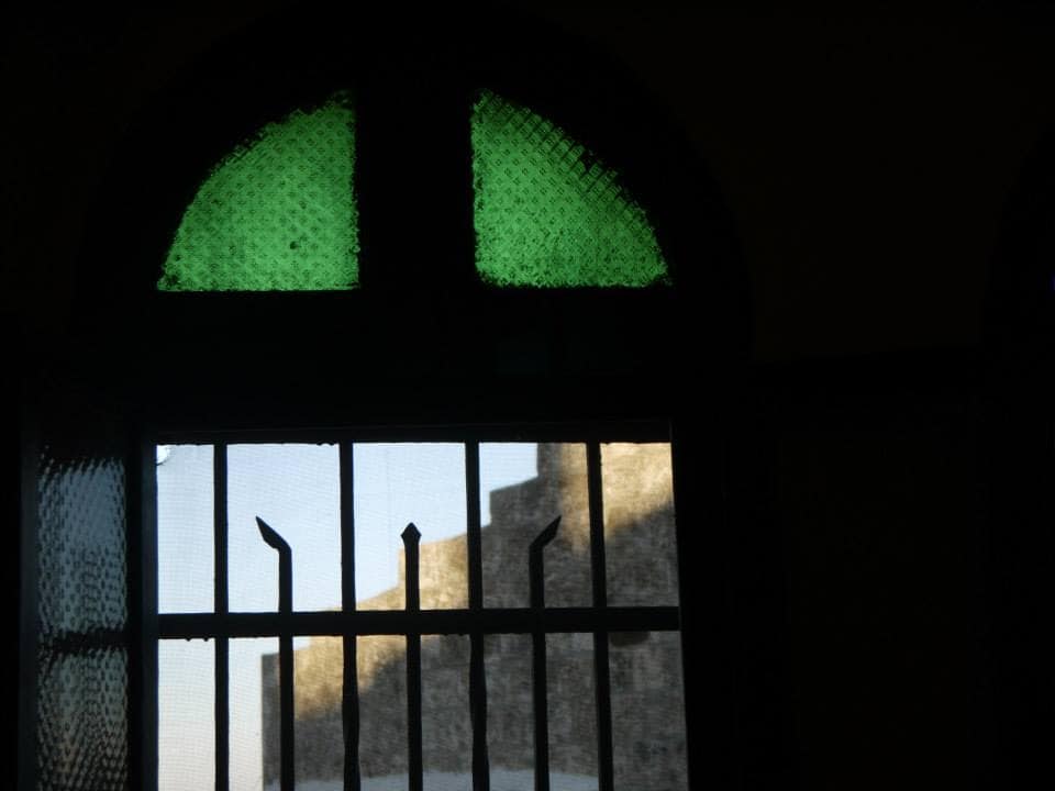 A photograph of an old stone building, taken looking out of a metal barred window with green stained glass panels at the top.