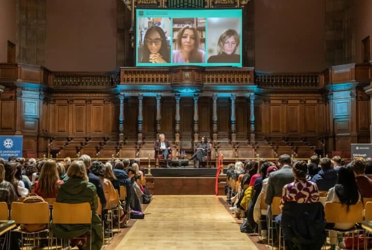 Photo taken from back of the audience facing a panel event with two in-person speakers and a screen above with virtual panellists