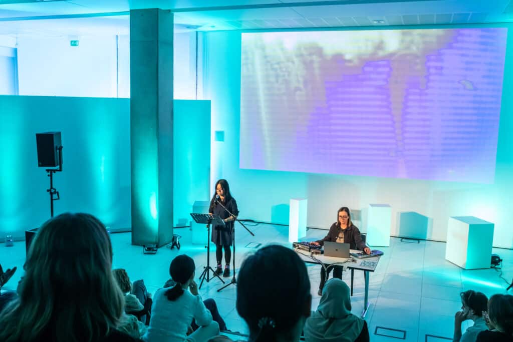 Two performers in an event - one is speaking in front of a microphone, another is playing a keyboard and sound machine