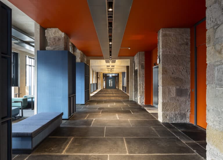 A modern hallway with an orange ceiling and stone walls features gray stone flooring. Blue cubicles and benches line the left side, while orange doors are on the right. Natural light streams in from windows on the left, illuminating the contemporary space.