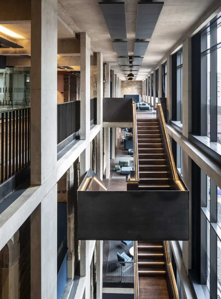 A modern, multi-level interior space featuring an industrial design with exposed concrete and large windows. A central staircase spans multiple floors, lit by recessed lighting. Glass walls and minimalist furniture add to the open, airy feel of the environment.