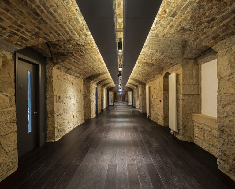 A long, narrow corridor with arched stone ceilings and walls, illuminated by modern ceiling lights. The polished dark wooden floor extends down the hallway, flanked by several doors on either side. The corridor exudes an old-world charm with a contemporary touch.