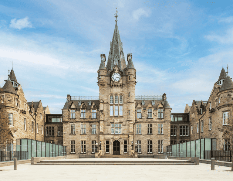 A grand historical building with a central tower featuring a large clock. The structure is symmetrically designed with multiple windows and pointed architectures on either side. It stands against a backdrop of a bright blue sky with some clouds.
