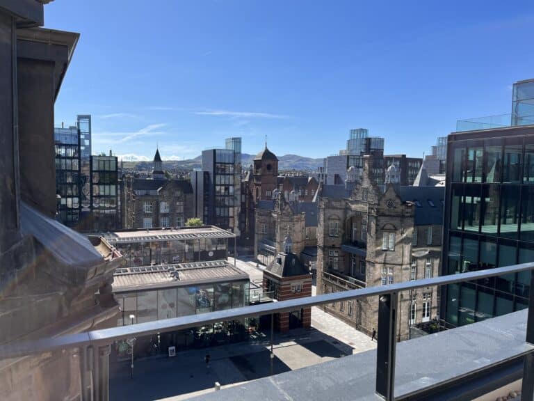 A view from a high balcony overlooking a cityscape with historical and modern buildings under a clear blue sky. A central square is visible below, surrounded by various architectural styles, with hills in the distant background.