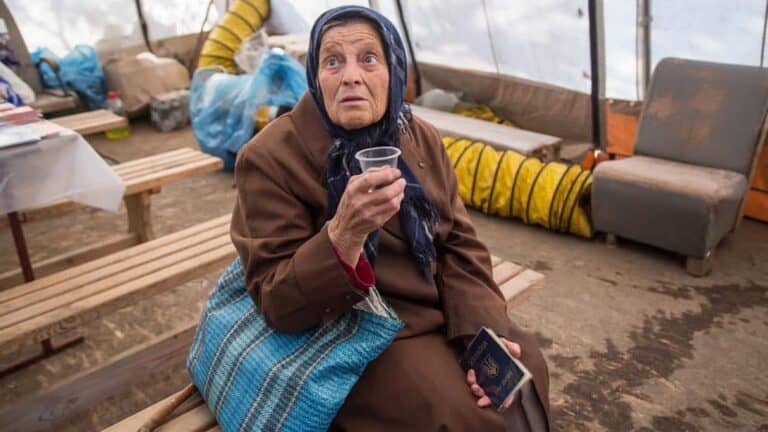 An elderly woman wearing a headscarf and brown coat sits on a wooden bench in a tent. She holds a small glass in one hand and her passport in the other. She has a blue striped bag next to her. The background shows packed items and yellow ventilation tubes.