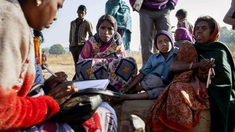 A group of women and a child in colorful clothing sit together outdoors on a sunny day. One woman is writing in a notebook, while the others look on attentively. In the background, more people stand and the scene appears to be set in a rural area.