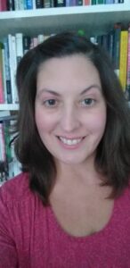 A woman with shoulder-length brown hair, wearing a red top, is smiling at the camera. She is standing in front of a white bookshelf filled with various books.
