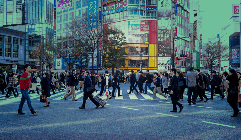 A bustling city intersection with many people crossing the street in various directions. Background includes tall buildings adorned with large billboards, signage, and storefronts. Some trees without leaves are also visible among the urban scenery.