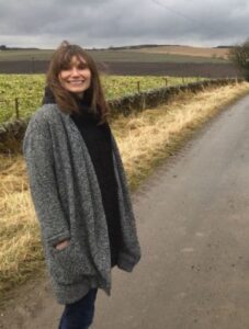 A person dressed in a grey coat, black shirt, and blue jeans stands beside a rural road. The background features a farm field with patches of grass and a cloudy sky. The person is smiling and has their hands in their coat pockets.