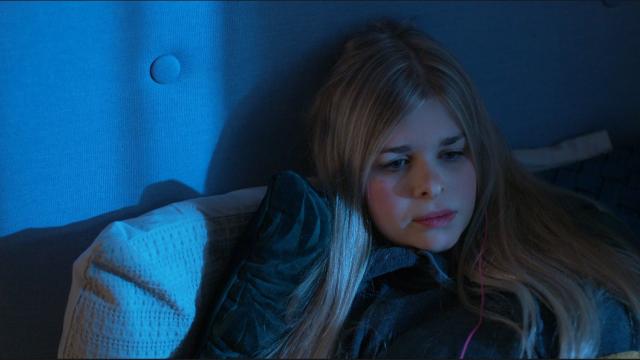 A young person with long, blonde hair lies on a pillow, wearing a dark shirt. They appear to be looking off to the side in deep thought. The lighting has a blueish tint, giving the scene a calm and reflective mood.