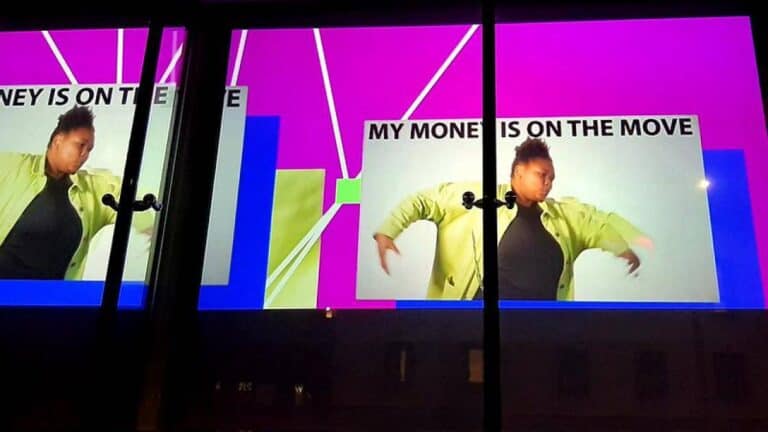 A digital billboard at night shows a vibrant advertisement featuring a person in a light green jacket against a bright pink and blue background. The text reads, "MY MONEY IS ON THE MOVE." The scene is reflected in glass in front of the billboard.