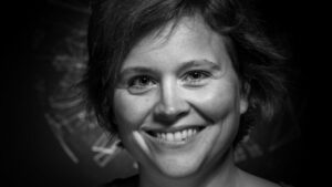 Close-up black-and-white photograph of a smiling person with short hair. The background is dark with faint lines and geometric shapes. The person is looking directly at the camera.
