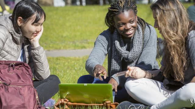Three young women sit on the grass, gathered around a green laptop. They are smiling and appear engaged in conversation. One woman points at the laptop screen, while another holds a phone. A red backpack rests nearby. The scene is set outdoors on a sunny day.