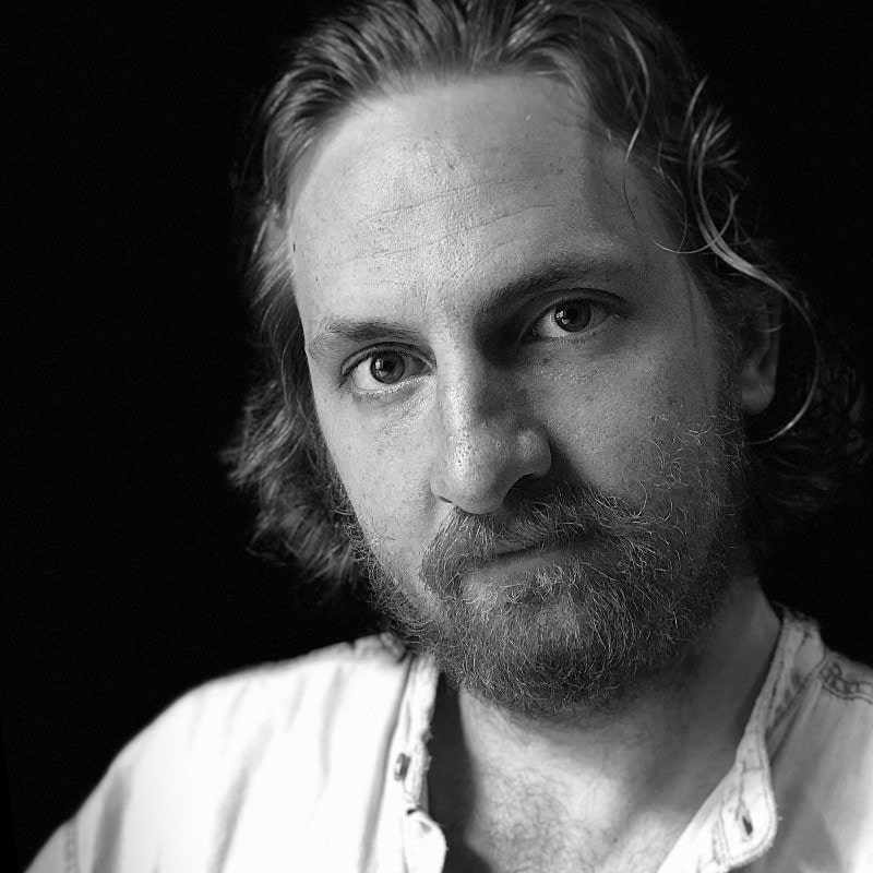 A grayscale portrait of a man with medium-length, wavy hair and a beard. He is wearing a light-colored shirt with a slightly open collar. The background is dark, making his face stand out prominently. The expression on his face is calm and contemplative.
