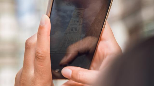 A person is holding a smartphone with a reflection of a tall, narrow building, resembling a historic or architectural structure, visible on the screen. The person's fingers are poised to interact with the phone. The background is blurred.