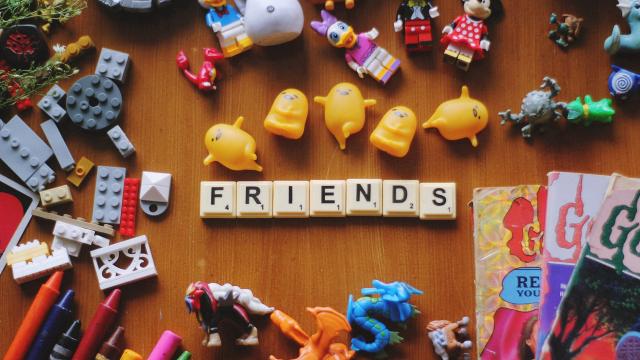 A collection of toys surrounds the word "FRIENDS" spelled out with Scrabble tiles on a wooden surface. Among the toys are action figures, crayons, building blocks, and miniature characters.