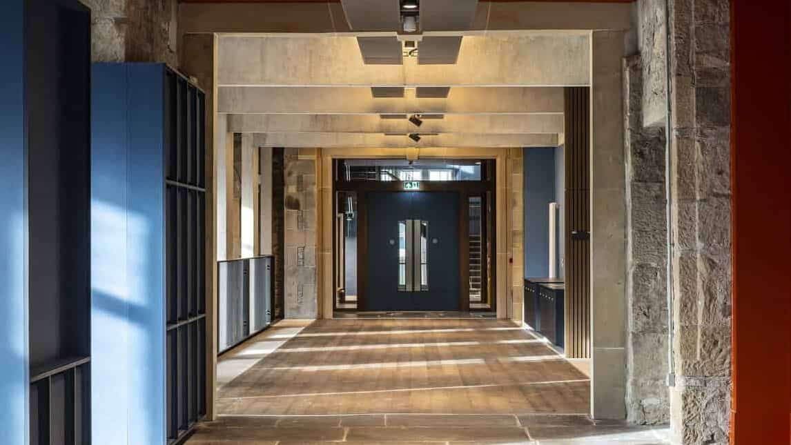 A modern corridor features stone walls, exposed ceiling beams, and wooden flooring illuminated by natural light. Blue and red wall sections add color, and a glass door at the end leads to another area. Geometric ceiling lights run the length of the hallway.