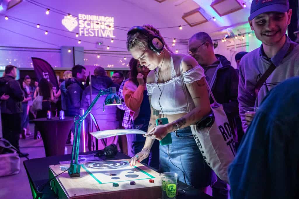 A young woman wearing headphones interacts with an exhibit at the Edinburgh Science Festival. She is standing at a table with various tools and materials, while other festival-goers are gathered around, engaging with different exhibits. Event signage is visible in the background.