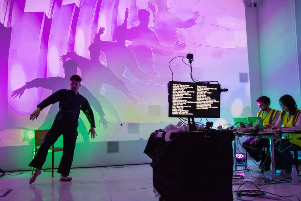 A performer dances in front of a colorful projection of shadowy figures, their movements being captured and displayed on a screen with code. Two people in high-visibility vests watch from a desk with equipment in the background.