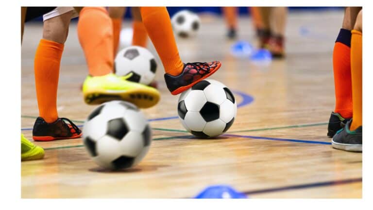 Children wearing bright orange socks and various colored shoes engage in a soccer practice indoors. Multiple soccer balls and blue training cones are placed on the wooden floor as the children dribble and move around the space.