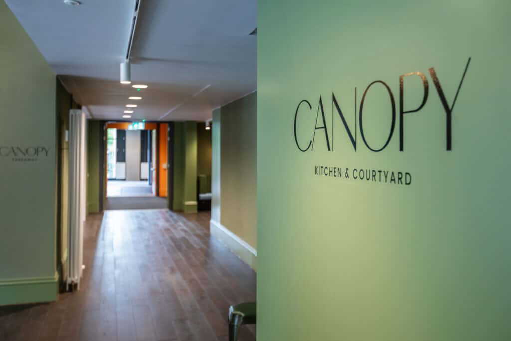 A brightly lit hallway leads to an open area with natural light streaming in. On the right wall, "CANOPY Kitchen & Courtyard" is prominently displayed in sleek, modern lettering. The hallway floor is wooden, and soft green tones adorn the walls.
