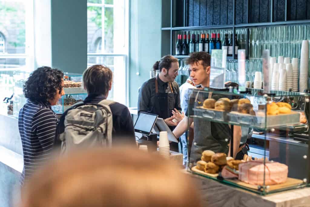 Customers wait in line at a modern cafe. Two baristas, one holding a tablet, are engaged in conversation with a customer. Shelves behind them are stocked with wine bottles, cups, and stacked baked goods. Natural light filters through large windows illuminating the space.