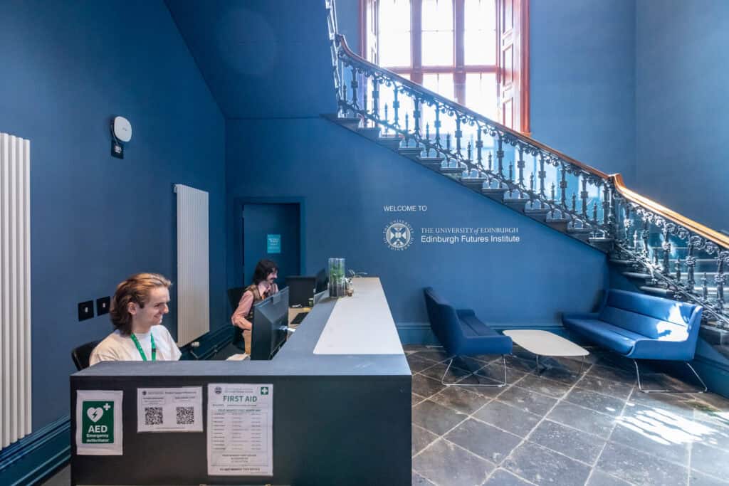 A reception area of The University of Edinburgh Futures Institute with two receptionists at a desk. There's a staircase with ornate railing leading to upper levels. Blue walls, a large window, and lounge chairs create a welcoming atmosphere. An AED device is visible.