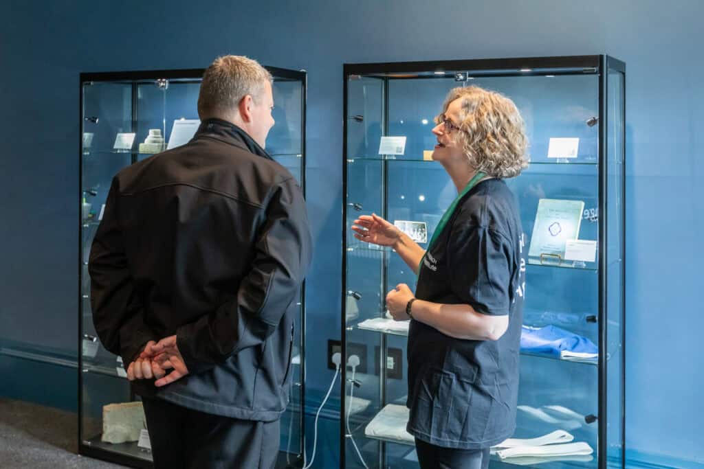 Two people are conversing in front of glass display cases showcasing various items. They are dressed in casual, dark clothing and appear to be in a museum or exhibition setting. Both individuals are engaged in a friendly discussion, with one gesturing animatedly.