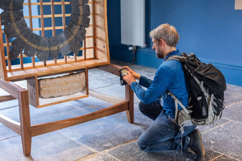 A man with a gray beard and a backpack kneels on one knee, photographing an exhibit in a museum. The exhibit features a large, circular artifact with inscriptions, displayed on a wooden stand. The background has a blue wall and tiled floor.