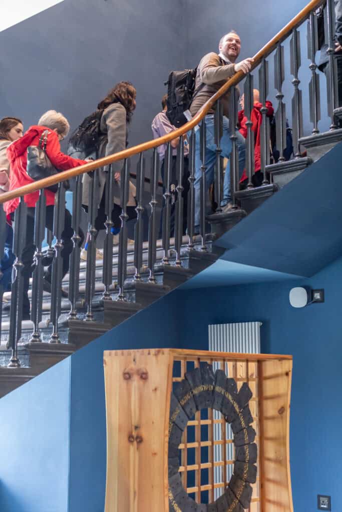 A group of people climb a wooden and metal staircase in a blue-walled building. Some carry backpacks, and they are ascending together. In the foreground, there is a wooden structure with round gear-like components.